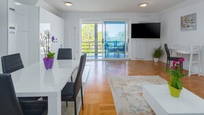 A modern living room with a white sofa, a coffee table, and a TV mounted on the wall. There is a dining area with a white table and chairs, and a balcony with a view of greenery. - Modern two bedrooms apartment Marea Blue with sea view in Funtana (Funtana)
