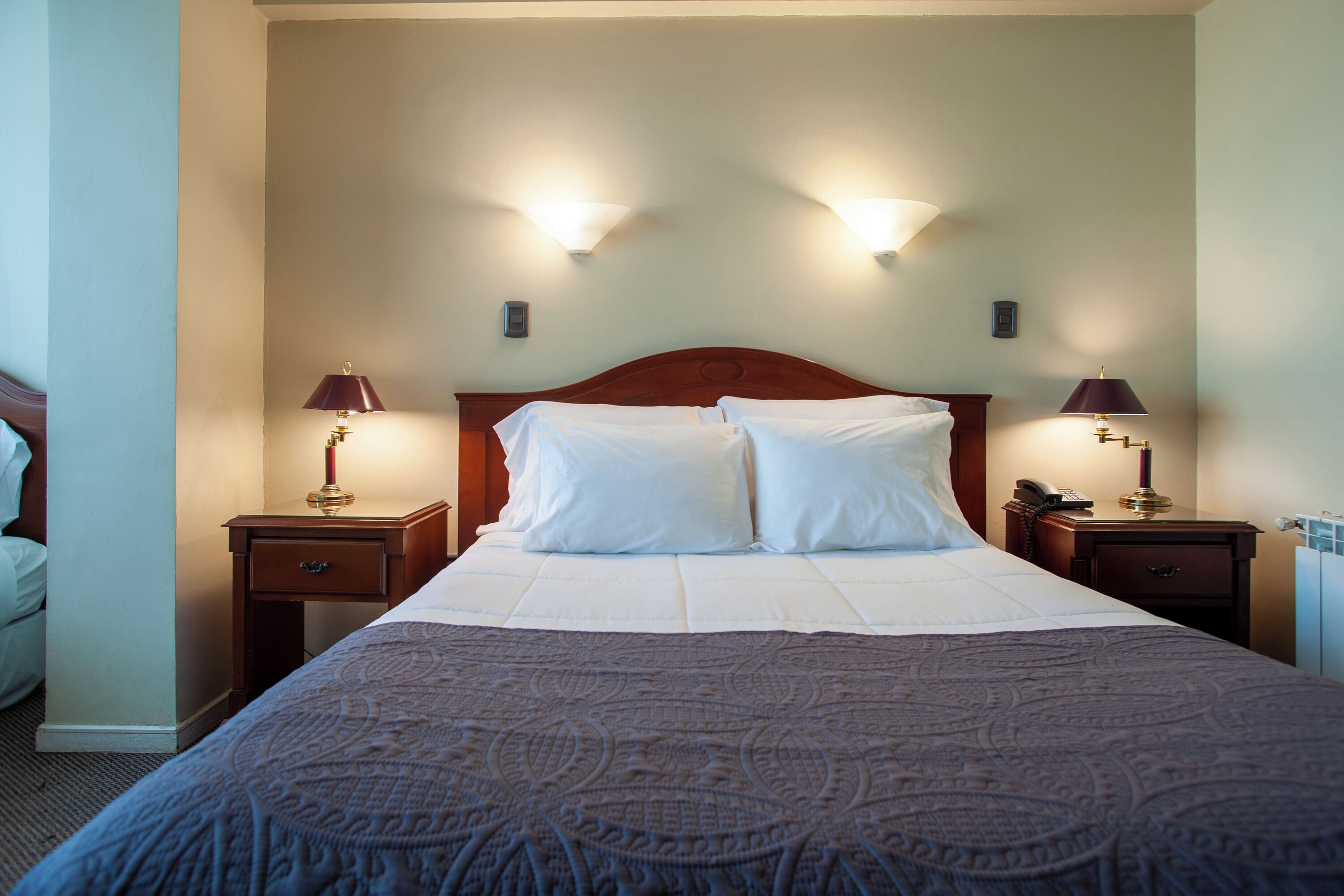 Una cama bien tendida con sábanas blancas y una colcha estampada, flanqueada por dos mesitas de noche con lámparas, en una habitación de diseño sencillo y limpio.