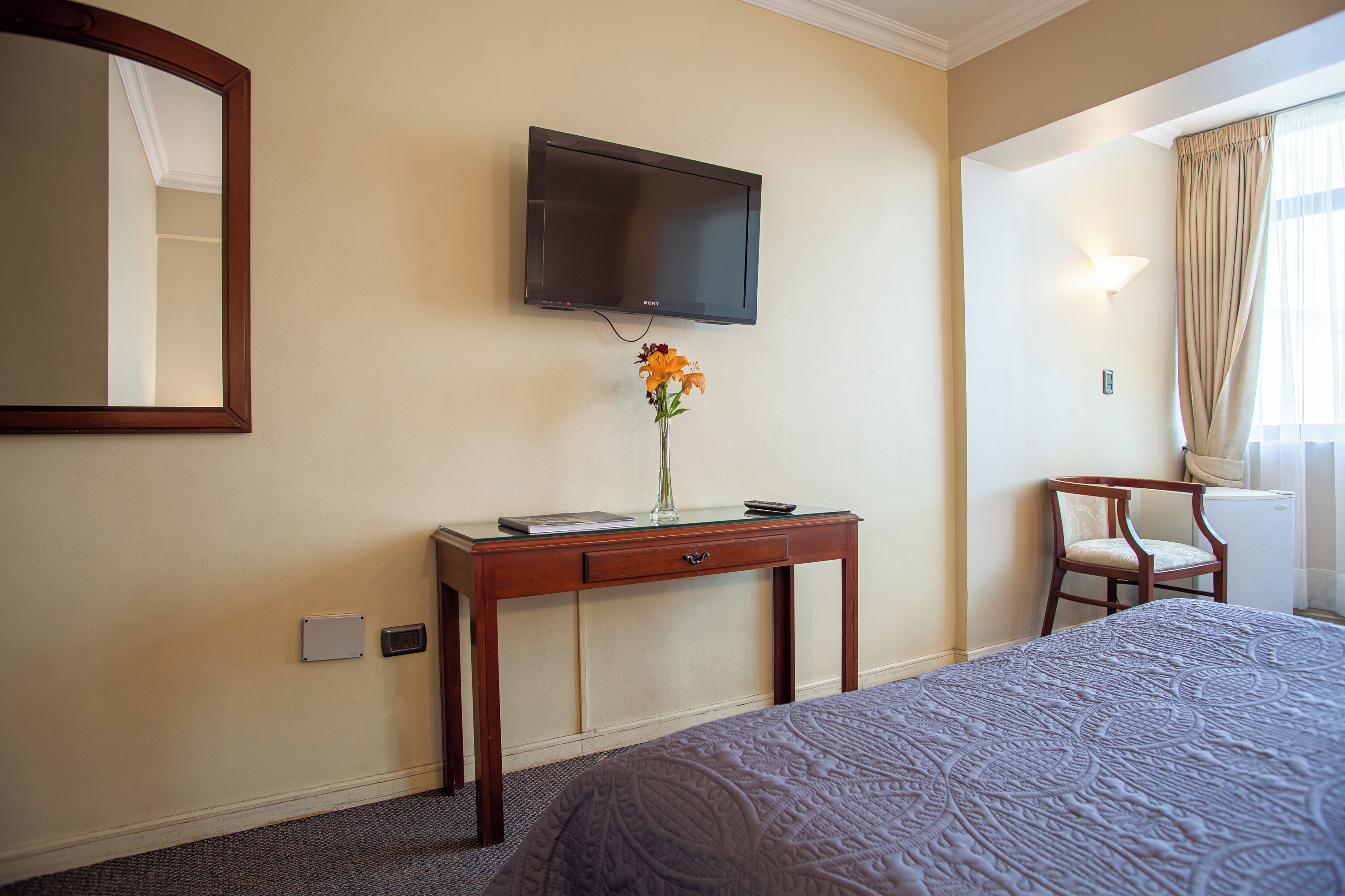A hotel room with a bed, a desk with a vase of flowers, a television mounted on the wall, and a chair.