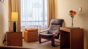 Een hotelkamer met een leren fauteuil, een klein voetenbankje, een nachtkastje met een lamp en een vaas met bloemen.