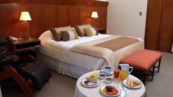Een hotelkamer met een net opgemaakt bed, een nachtkastje met een lamp, een stoel, en een ontbijttafel met borden eten en drinken.