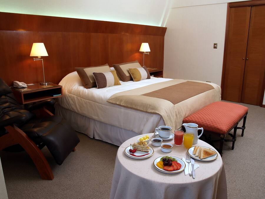 Een hotelkamer met een net opgemaakt bed, een nachtkastje met een lamp, een stoel, en een ontbijttafel met borden eten en drinken.