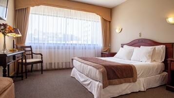 Ett hotellrum med en stor säng, ett skrivbord med en lampa, en stol och ett fönster med gardiner.