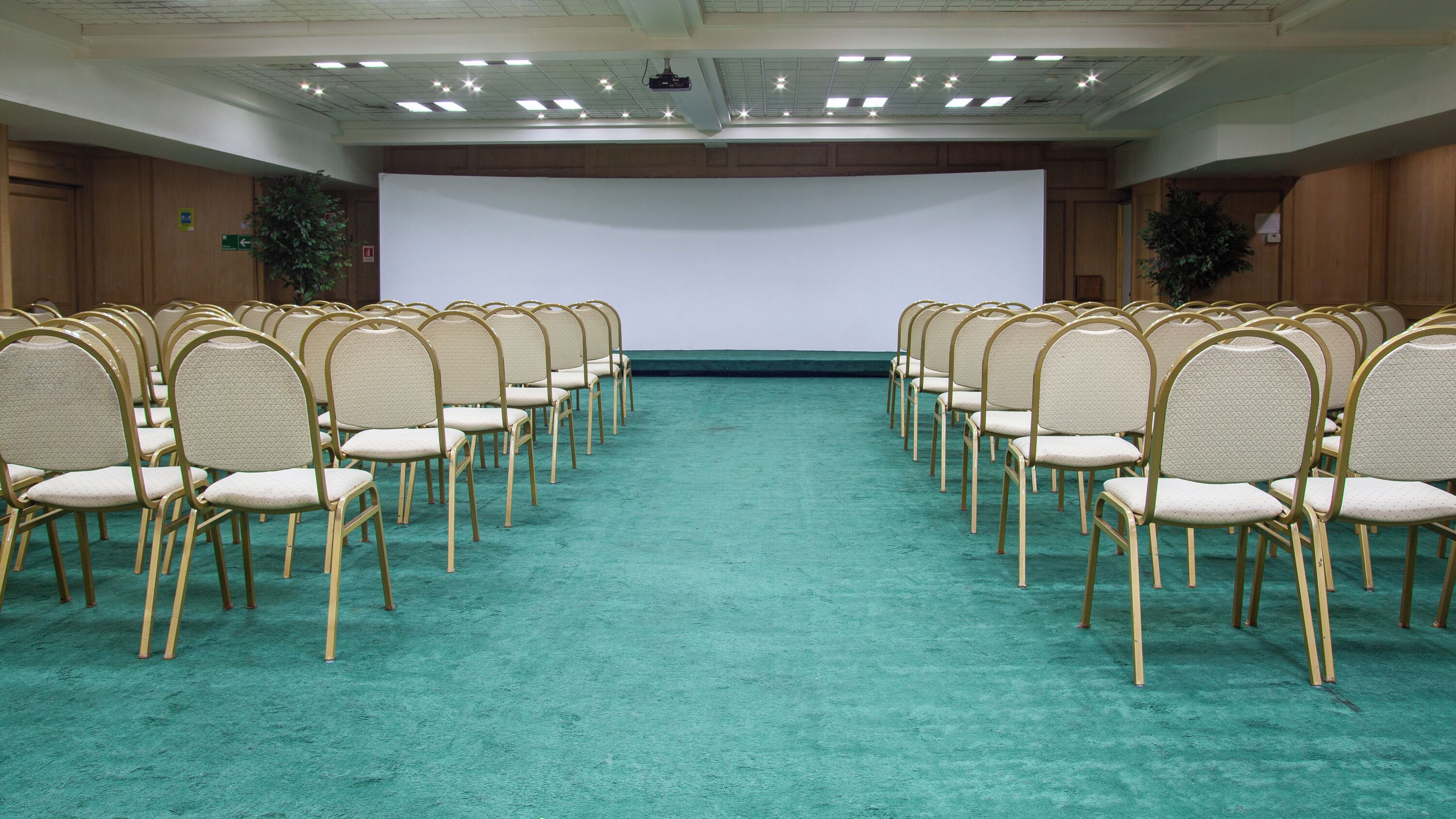 A conference room with rows of chairs facing a large white screen, carpeted floor, wooden panel walls, and ceiling lights.