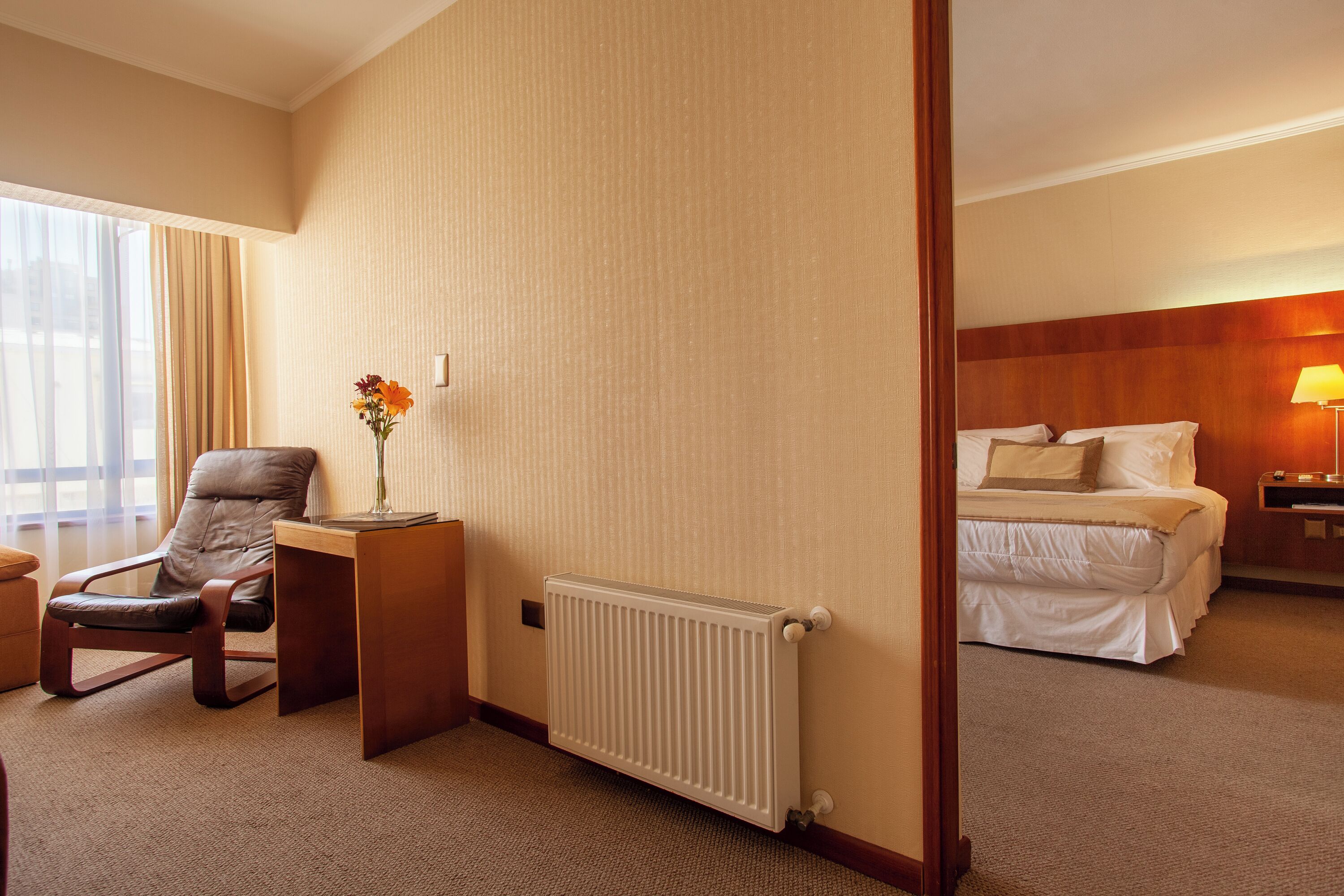 Una habitación de hotel con una cama, un escritorio con una silla, un radiador y un florero con flores.