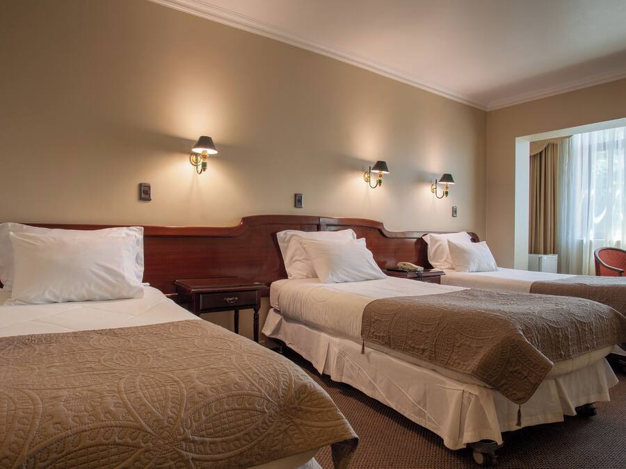A hotel room with three beds, each with white bedding and a brown throw blanket. There are wall-mounted lamps on either side of the beds.