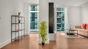 A modern living room with a wooden floor, a black sofa, a coffee table, and a tall bamboo plant. - Stunning Serviced Apartment-Canary Wharf (London)
