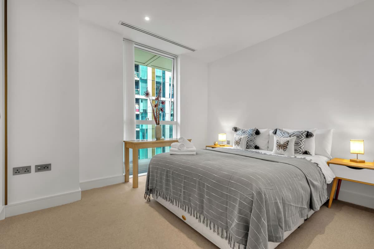 Ein modernes Schlafzimmer mit einem großen Bett, Nachttischen, einem Fenster mit Aussicht und einem kleinen Tisch mit einer Vase.