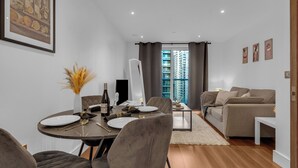 A modern living room with a dining area, featuring a sofa, a coffee table, and a dining table set for four. - Stunning Serviced Apartment-Canary Wharf (London)