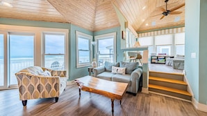 A spacious living room with a wooden ceiling, large windows, a sofa, a coffee table, and a staircase leading to an upper level.