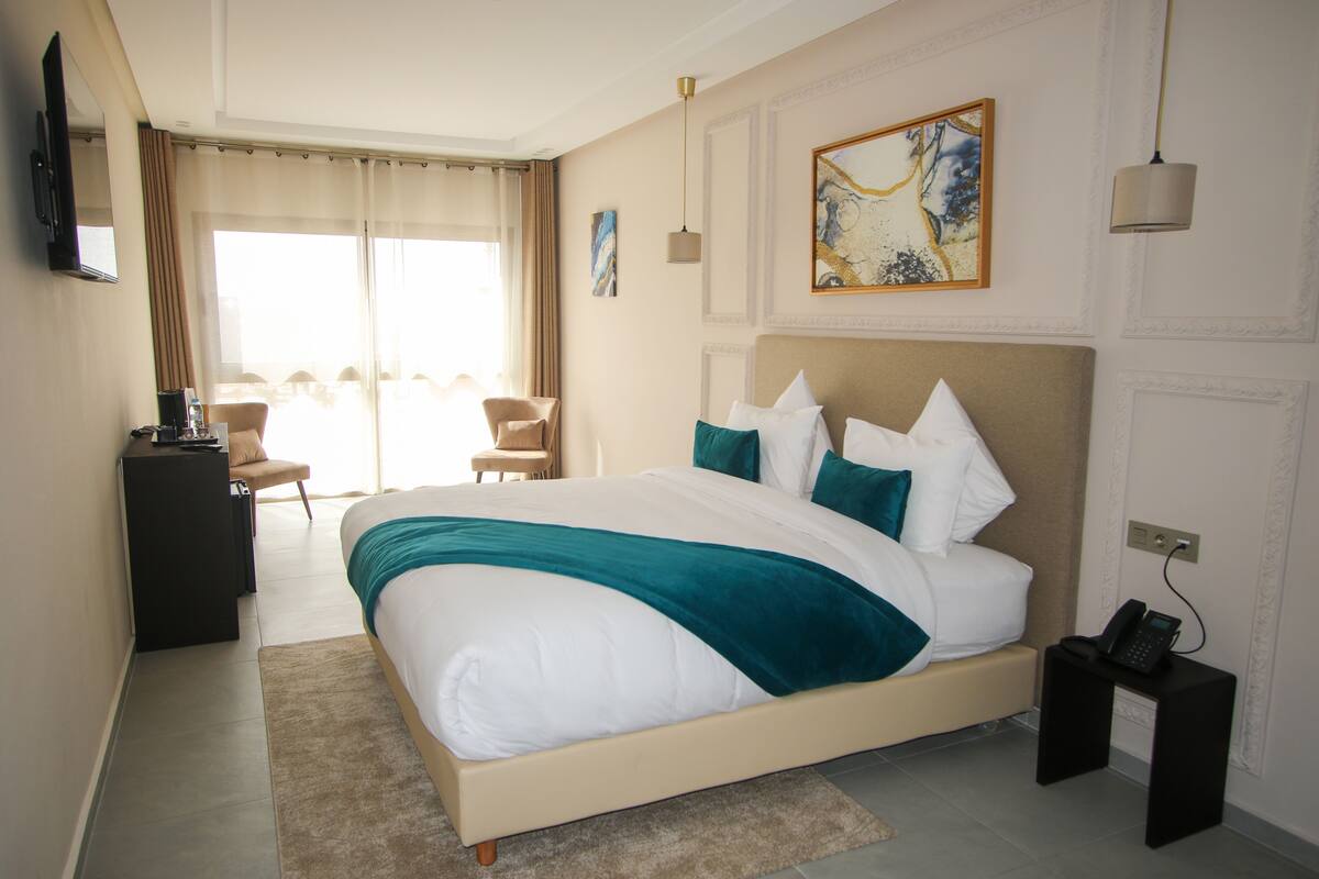 Ein Hotelzimmer mit einem großen Bett, einem Schreibtisch mit Computer, einem Sessel und Blick ins Freie.