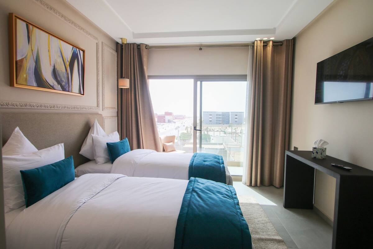 Ein Hotelzimmer mit zwei Betten, einem Fernseher, einem Schreibtisch und einem Balkon mit Vorhängen.