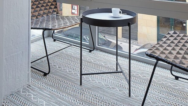A balcony with patterned floor tiles, a wicker chair, a small round table with a cup, and a view of a building with balconies and a sign reading "ESTAURANTE". - Azoka Suite by FeelFree Rentals (Donostia)