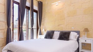 Una cama bien tendida con ropa de cama blanca y gris, una mesita de noche con una lámpara y una pared de piedra.