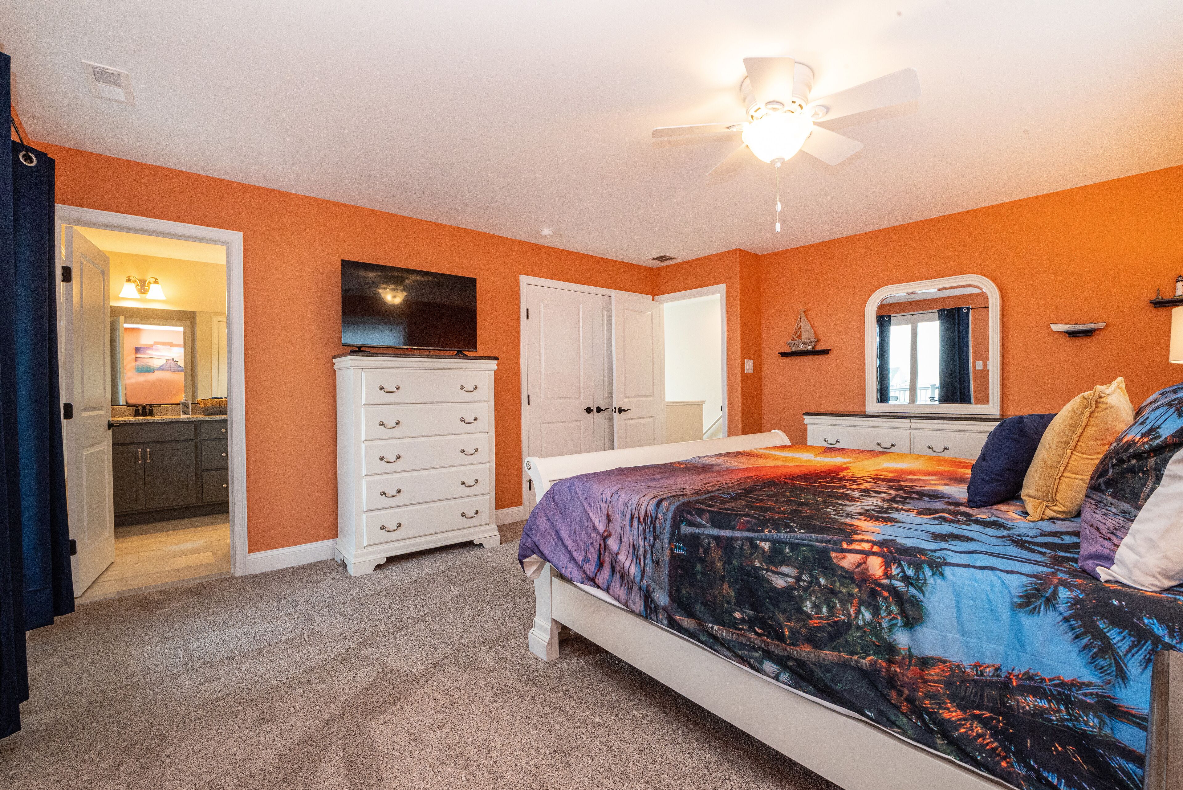 A bedroom with an orange wall, a bed with a tropical print bedspread, a ceiling fan, and a flat-screen TV mounted above the dresser.