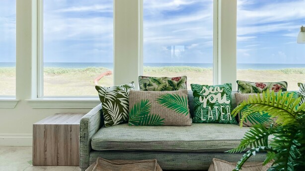 Een kamer met een groot raam met uitzicht op een strand, een groene bank met tropische kussens, en een houten bankje.