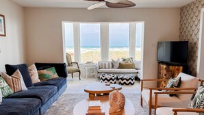 Een woonkamer met een donkerblauwe bank, een houten salontafel en een beige fauteuil. Er is een televisie op een houten kast en een groot raam met uitzicht op het strand.