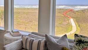 Een kamer met een groot raam dat uitkijkt over een kustlandschap, met een pad en een rode constructie.