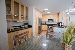 A modern kitchen with wooden cabinets, stainless steel appliances, and a glass dining table. - Book Chillaxin 214B | Sandbridge Vacation Rental | Ocean Sands Realty (Virginia Beach)
