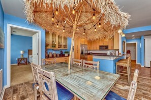 A dining area with a glass table, wooden chairs, and a thatched ceiling with hanging lights. - Book Chillaxin 214B | Sandbridge Vacation Rental | Ocean Sands Realty (Virginia Beach)