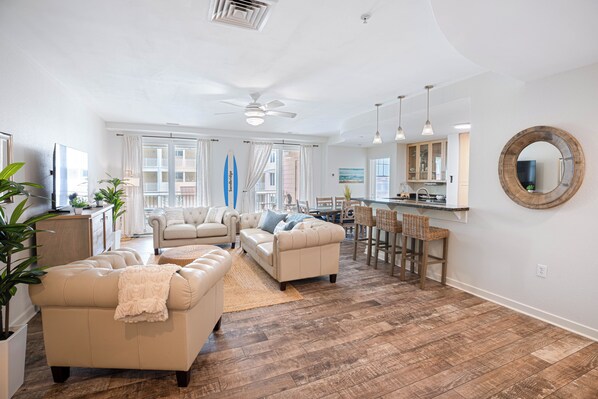 A spacious living area with a sofa set, a dining area with bar stools, and a kitchen in the background.
