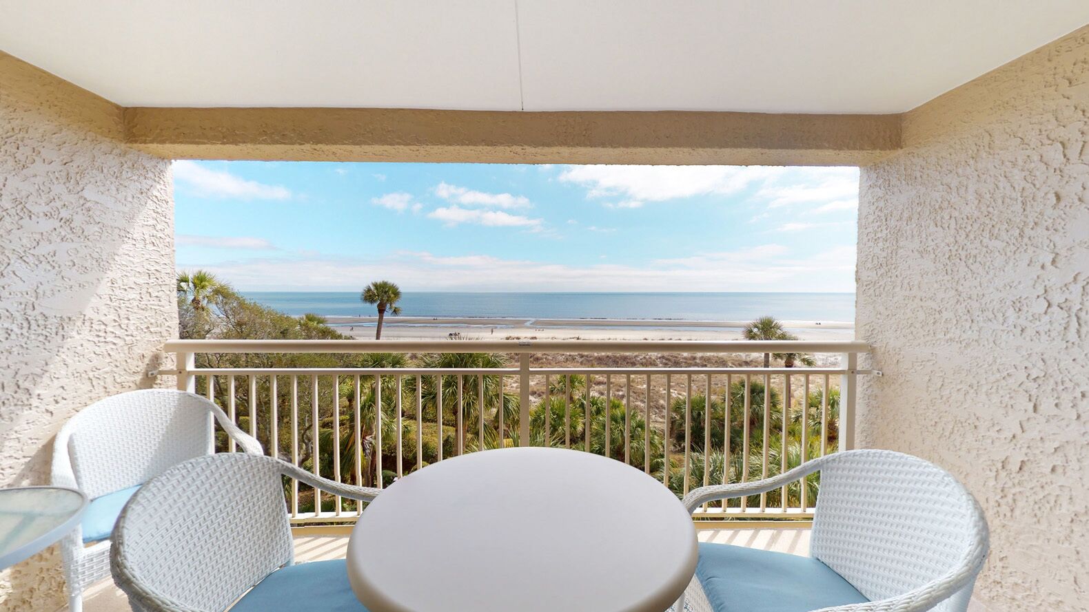 A balcony with a view of the beach, white wicker furniture, and a small table.