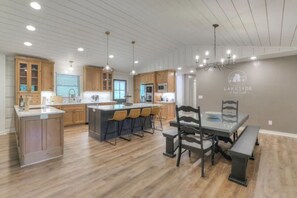 A modern kitchen with wooden cabinets, a central island with bar stools, pendant lighting, and a dining area with a bench and table. - Lakeside Family Escape-Modern log home in secluded cove w/pvt. dock, Sleeps 12 (Lynchburg)