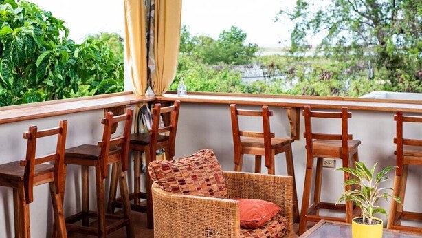 Een zithoek met rieten stoelen en een tafeltje, een potplant en uitzicht op groen en water.