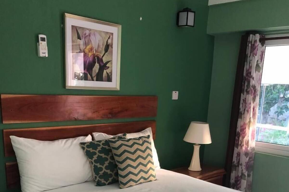 A neatly made bed with a wooden headboard, a framed picture on the wall, a lamp on the nightstand, and a window with curtains.
