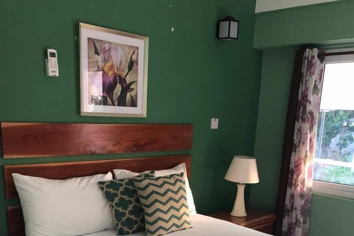 A neatly made bed with white bedding and two patterned pillows, a wooden headboard, a framed picture on the wall, a bedside lamp, and a window with curtains.