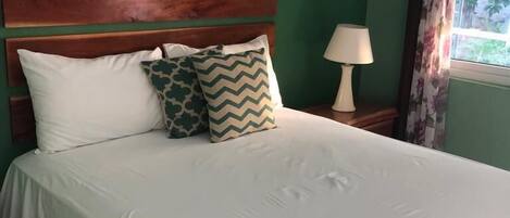 A neatly made bed with white bedding and two patterned pillows, a wooden headboard, a framed picture on the wall, a bedside lamp, and a window with curtains.