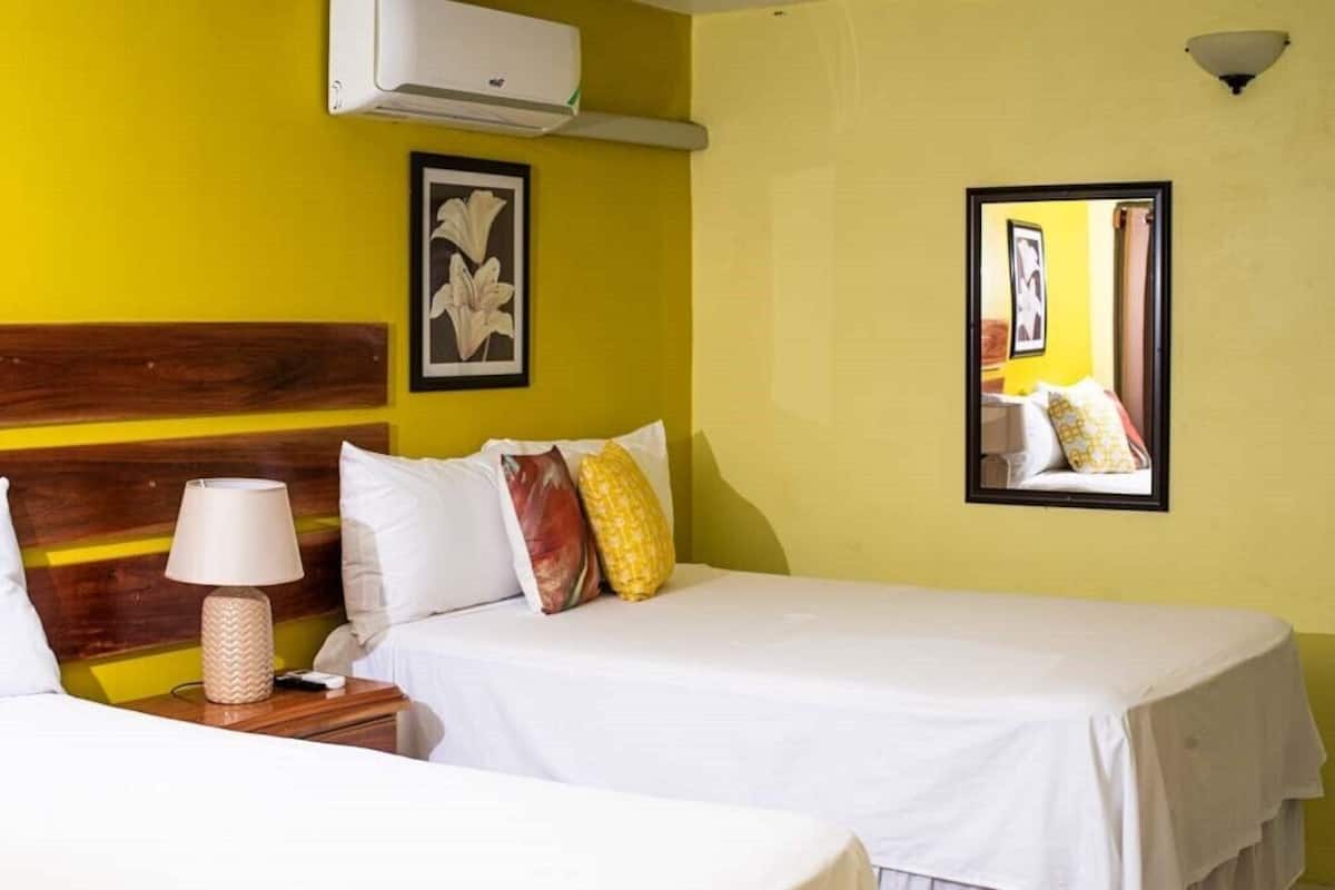 A hotel room with two beds, a wooden headboard, a wall-mounted air conditioner, a framed picture, and a bedside lamp.