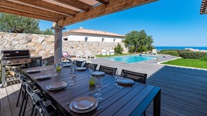 Un área de comedor al aire libre con vista a una piscina, pared de piedra y una casa al fondo.