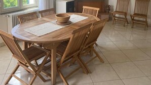 Ein Essbereich mit einem runden Holztisch, sechs Holzsesseln und einer Holzküche auf dem Tisch. Ein Fenster mit Vorhängen, eine weiße Klimaanlage und eine Wand mit drei gerahmten Bildern sind zu sehen.