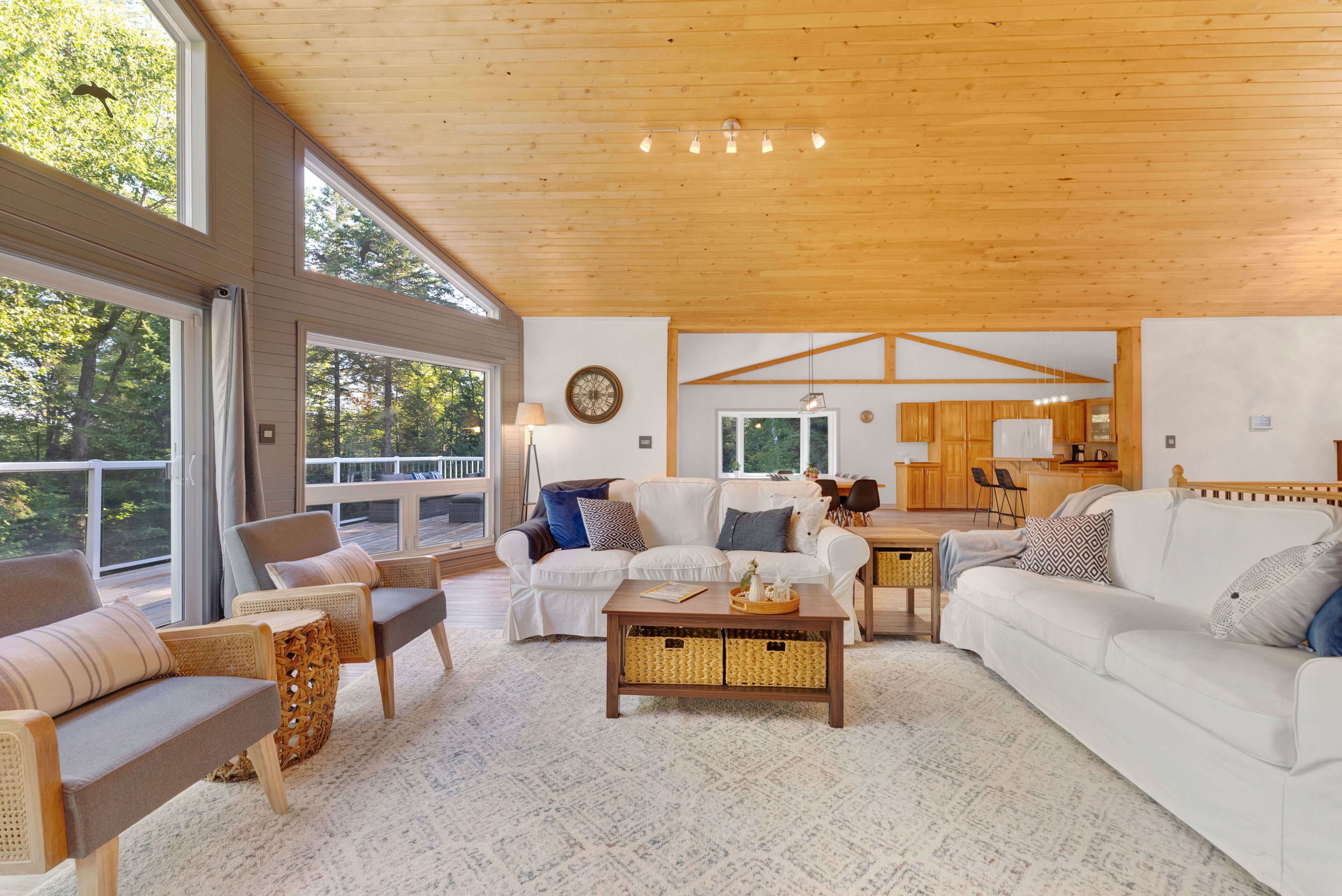 A spacious living room with a large white sofa, a wooden coffee table, and wicker side tables. There is a kitchen area in the background with wooden cabinets and a dining table.