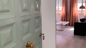 Habitació amb una porta verdosa, paret blanca, sòl de fusta, sofà negre, taula blanca i finestra amb cortines.