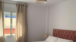 Un dormitori amb un llit de fusta, roba de llit blanca, un panell de capçal tapissat de color rosa, una làmpada al sostre i una finestra amb cortines.