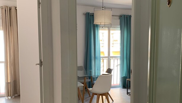 A room with a dining area, a door with a golden handle, and a view through a window with curtains. - Terraza Catedral Olehousing 6 (Sevilla)