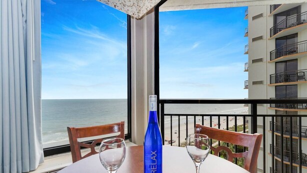 A balcony with a view of the ocean, a bottle of RELAX BIESLING, and two empty wine glasses on a wooden table. - Impressive 1Br Oceanfront/Dogs OK! (Myrtle Beach)