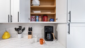 Une cuisine avec des meubles blancs, un plan de travail en marbre, une machine à café, une courgette jaune, un bloc à couteaux et une tasse.