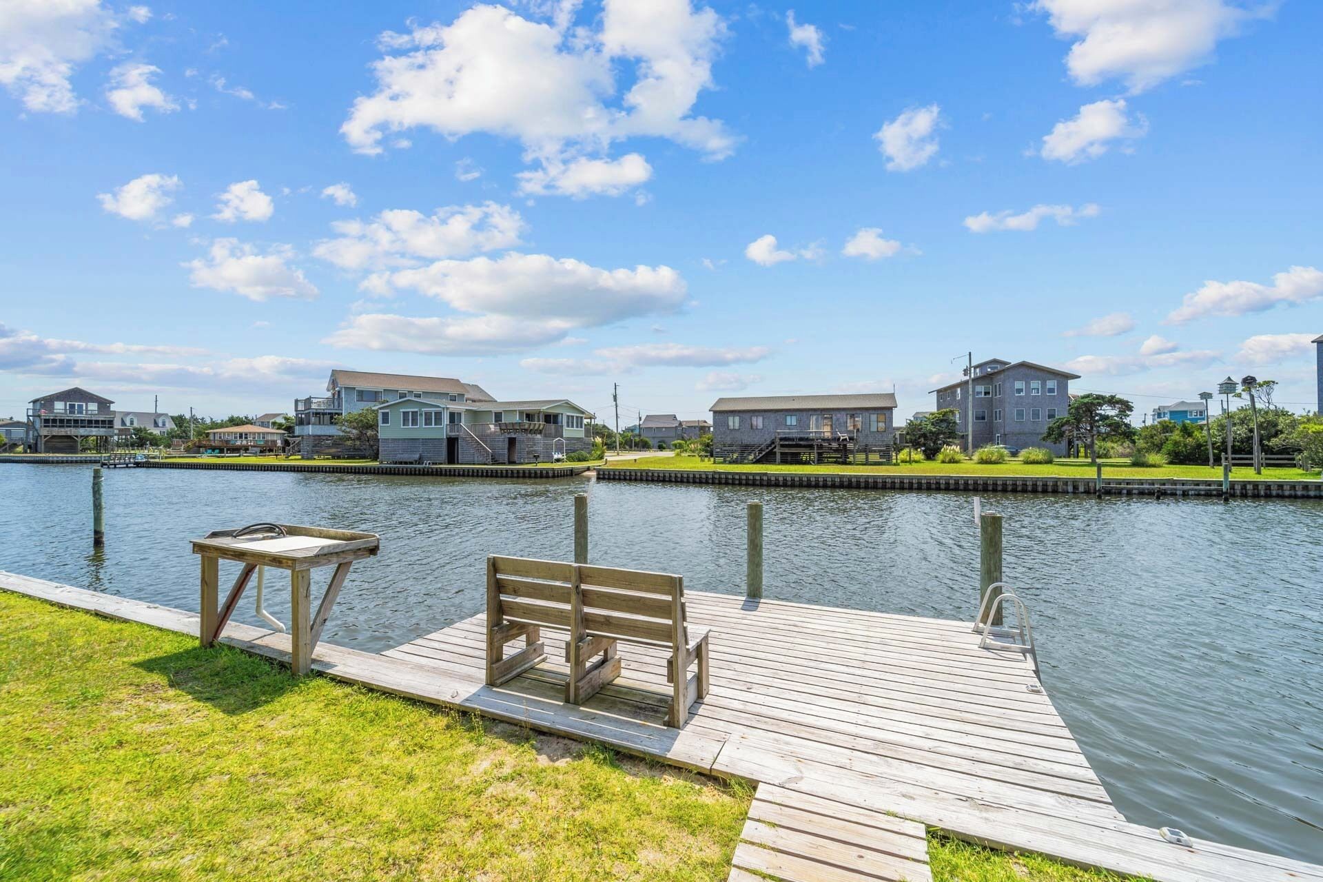 A wooden dock with a bench and a table, leading to a calm water body with houses in the background.
