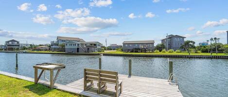 A wooden dock with a bench and a table, leading to a calm water body with houses in the background.