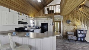 A spacious kitchen with white cabinets, a black refrigerator, and a large island with bar stools. There is a wooden ceiling with exposed beams and a clock on the wall. - Canalfront Dog Friendly w/ Hot Tub, Boat Dock, Game Room, Nearby Beach Access (Avon)