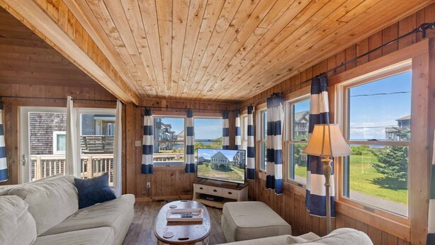 A cozy cabin interior with a wooden ceiling, a large window with a view of a house and greenery, a sofa, a coffee table, and a lamp. - Canalfront Dog Friendly w/ Hot Tub, Boat Dock, Game Room, Nearby Beach Access (Avon)