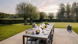 An outdoor dining area with a long table set for a meal, surrounded by a well-manicured lawn and trees.
