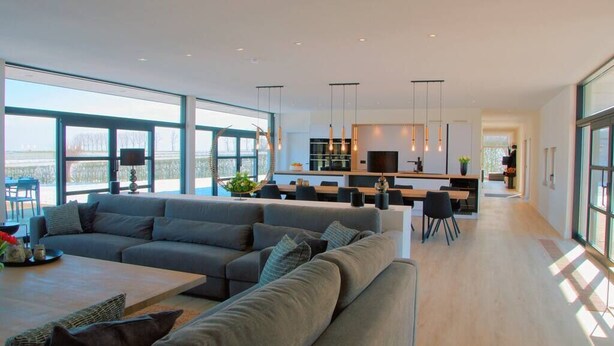 A spacious living area with a large grey sectional sofa, a wooden coffee table, and a dining area with a kitchen in the background.