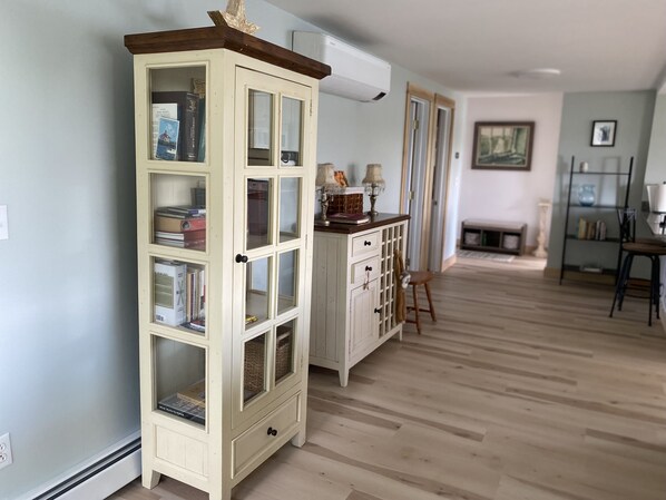 Una habitación con un mueble alto y estrecho con varios estantes, un banco de madera, una mesita con una lámpara y una chimenea al fondo.