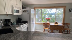 Una cocina con gabinetes blancos, encimera de granito, estufa, microondas y refrigerador. Se ve una ventana con vistas a árboles y una mesa con sillas.