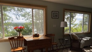Un comedor con una mesa y sillas de madera, un jarrón con flores y vistas a árboles a través de amplias ventanas.