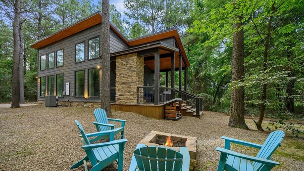 A modern cabin with a stone foundation, large windows, and a deck. There are Adirondack chairs and a fire pit in the foreground.